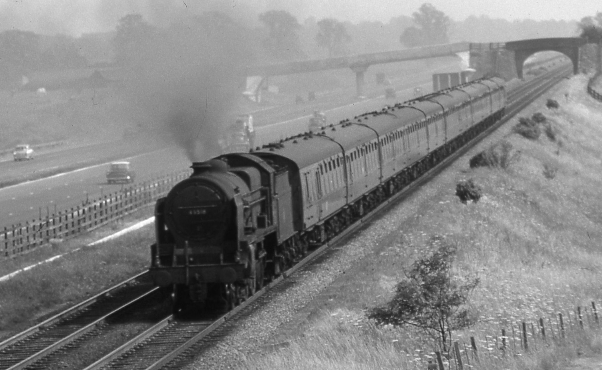 Un-rebuilt and un-named Patriot 45510 on a Euston-Manchester express near Watford Gap with infant M1 in the background in June 1960. – Derek Cross A large steam locomotive hauls an express passenger train along a low, straight embankment in open countryside, adjacent to the recently-built M1 motorway.